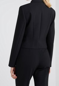 Blazer nero su misura con una vestibilità slim, caratterizzato da spalle strutturate, colletto a rotondo e dettagli a pieghe sul retro.