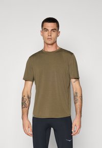 Homem com cabelo curto e escuro, vestindo uma t-shirt verde-oliva e leggings pretos, de pé contra um fundo claro e liso.
