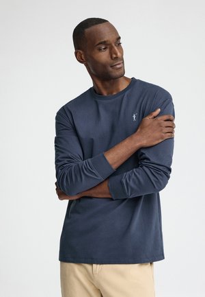 Mand iført navyblå langærmet skjorte og beige bukser, stående med armene krydset, kigger mod venstre mod en ensfarvet hvid baggrund.