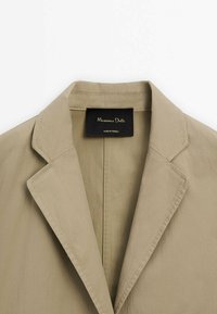 Beżowa marynarka z klapą z cięciem w szpic, wykonana z gładkiej tkaniny. Posiada subtelne przeszycia oraz czarną metkę z białym napisem wewnątrz.