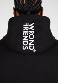 Zwarte hoodie met een grote witte print op de rug waarop "WRONG FRIENDS" staat. Heeft een comfortabele, casual uitstraling en een zachte textuur.