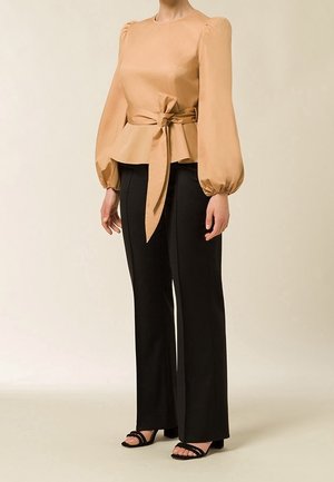 Kobieta w beżowej bluzce z długim rękawem i wiązaniem w talii, czarne spodnie o prostych nogawkach oraz czarne czółenka z odkrytymi palcami, stojąca na jednolitym tle.