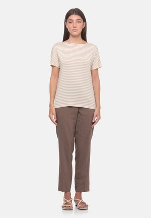 Donna in piedi che indossa un top beige a righe a maniche corte, pantaloni marroni dritti e sandali beige con le dita scoperte su uno sfondo semplice.