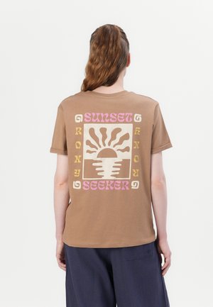 Osoba o długich włosach stoi tyłem, ubrana w brązowy t-shirt z stylizowanym słońcem i napisem „SUNSET SEEKER” w różowym i żółtym na plecach.