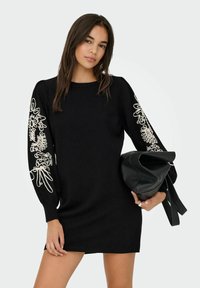 Zwarte gebreide jurk met witte bloemenborduursels op de gezwollen mouwen, ronde halslijn en geribbelde zoom. Model houdt een zwarte handtas vast.