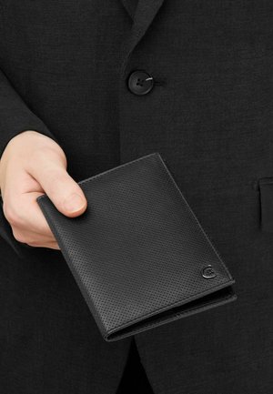 Mano sujetando una cartera de cuero negro texturizado contra una chaqueta de traje oscura con un botón visible.