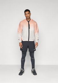 Lettvekts, fargeblokket jakke i rosa og hvitt, kombinert med svarte shorts og mønstrete strømpebukser. Har glidelås og hette.