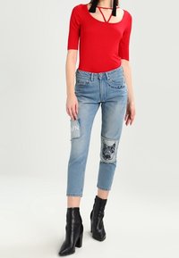 Body rojo ajustado con mangas cortas, combinado con jeans de color azul claro que presentan bolsillos de parche y diseños bordados, y botas de tacón negro hasta el tobillo.