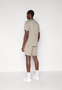 Άνδρας μοντέλο φοράει μπεζ αθλητικό t-shirt και κοντό παντελόνι που ταιριάζουν, συνδυασμένα με λευκά αθλητικά παπούτσια και κάλτσες. Διαθέτει απλό, λειτουργικό σχέδιο.