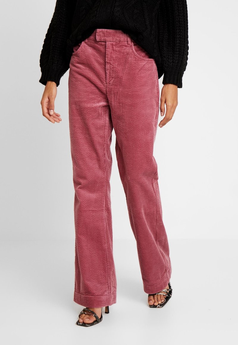 Femme portant un pantalon large en velours côtelé rose taille haute et un pull noir en maille, associé à des sandales noires à talons hauts et à brides.