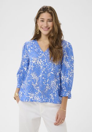 Jeune femme souriante portant un chemisier bleu à fleurs avec col en V et manches trois-quarts, ainsi qu'un pantalon blanc, accessoirisée de bijoux en or.