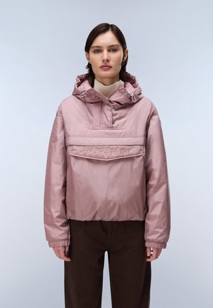 Une femme porte une veste à enfiler à capuche rose poudré, avec une poche à l’avant et un logo, associée à un pantalon foncé sur un fond uni.