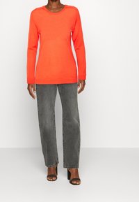 Pull à manches longues, col rond en orange vif, avec des poignets et un ourlet côtelés. Associé à un jean gris ample et des sandales noires à talons.