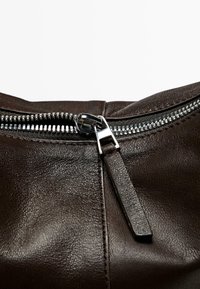 Gros plan sur la fermeture éclair d'un sac en cuir brun foncé partiellement ouverte avec un curseur en métal et une tirette en cuir assortie.