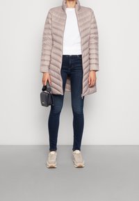 Ljusgrå puffjacka med horisontellt quiltat mönster, hög krage och öppen front. Bärs över en vit topp och mörka jeans, ihop med sneakers.