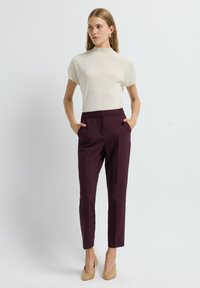 Vrouw in een beige gestructureerd T-shirt met korte mouwen, hoge paars op maat gemaakte broek en beige hakken, staand met handen in de zakken.