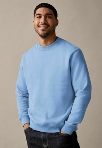 Lichtblauwe crewneck sweatshirt van zacht materiaal met ribbelboorden en -zoom. Model staat met handen in de zakken, draagt een donkere spijkerbroek.