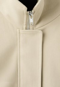 Gros plan du col d'une veste en tissu beige avec une fermeture éclair argentée partiellement fermée et un rabat rectangulaire cousu.
