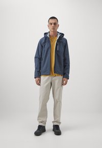 Marine blå jakke med hætte, grå foring, frontlynt og lommer, båret over en gul shirt, sammen med lyse beige bukser og sorte sko.