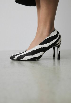 Woman wearing black and white zebra-striped high-heeled shoes standing on a smooth, light gray floor.
