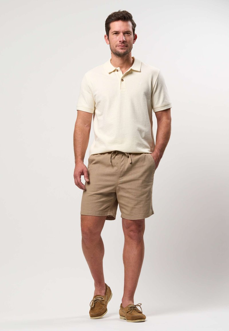 Homme debout portant un polo beige clair, un short beige et des mocassins marron sur fond blanc uni.