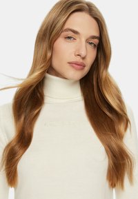 Maglione crema con collo a dolcevita realizzato in tessuto a coste, con logo in rilievo "GUESS JEANS" sul petto e lunghi capelli che cadono sulle spalle.