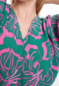 Detalle de una blusa con estampado floral en verde y rosa, con escote en V, llevada por una persona de piel clara y cabello castaño claro.