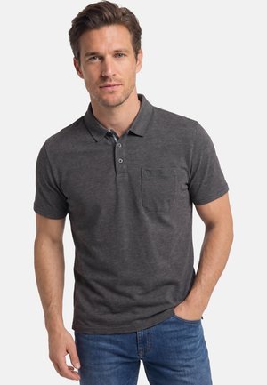 Mand iført mørkegrå kortærmet polo med brystlomme og blå jeans, stående med den ene hånd i lommen mod hvid baggrund.