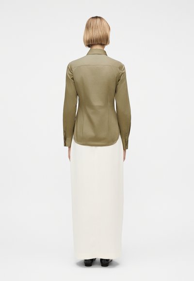 Άτομο με κοντά ξανθά μαλλιά, φοράει μια μακρυμάνικη μπλούζα χακί και μια λευκή μακριά φούστα, όρθιο με την πλάτη προς το μέρος μας σε απλό λευκό φόντο.