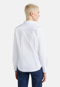 Camicia bianca a maniche lunghe con colletto strutturato, texture liscia e orlo curvo. Presenta polsini con bottone e un design a silhouette aderente.