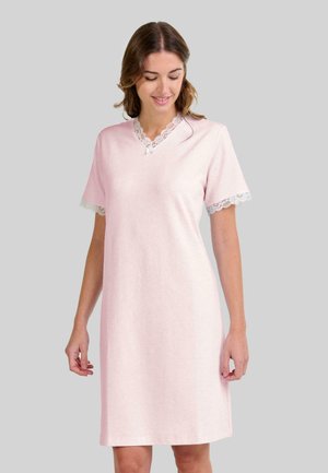 Mujer con un vestido de tiro a la rodilla de color rosa claro, con detalles de encaje en el escote y las mangas, de pie frente a un fondo gris liso.