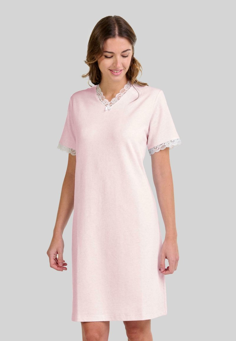 Mujer con un vestido de tiro a la rodilla de color rosa claro, con detalles de encaje en el escote y las mangas, de pie frente a un fondo gris liso.