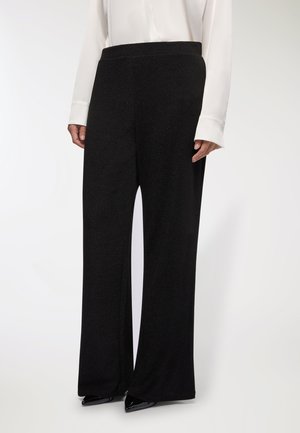 Pantaloni neri a gamba larga con una finitura testurizzata, caratterizzati da una vita alta e una silhouette fluida, abbinati a una blusa bianca a maniche lunghe.