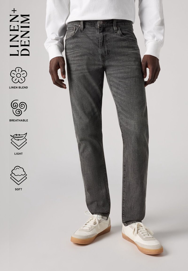 Homme portant un jean en denim gris mélangé lin et des baskets blanches, debout devant un fond clair uni. Le texte met en avant le lin, la respirabilité, la légèreté et la douceur.
