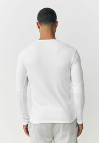 White long-sleeve ribbed shirt with a crew neckline, fitted design, and smooth texture, worn with light gray pants.
