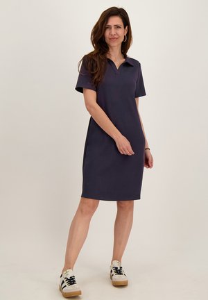 Vrouw met lang bruin haar, een donkerblauwe jurk met korte mouwen tot op de knie en witte sneakers die staat tegen een effen achtergrond.