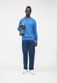 Moški v modrem puloverju, ohlapnih modrih kavbojkah, belih supergah in z sivo torbico čez ramo, stoji pred belim ozadjem.