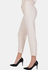 Donna che indossa pantaloni su misura di color crema e tacchi alti in pelle lucida nera, in posizione laterale su uno sfondo bianco.