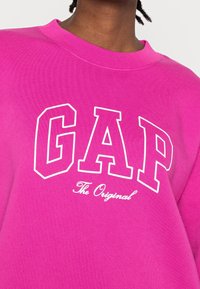 Felpa rosa con il logo "GAP" in lettere bianche in grassetto e il testo "The Original" sottostante in un carattere corsivo. Tessuto di cotone con finitura testurizzata.