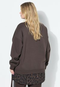 Mujer rubia con cabello ondulado de pie de espaldas, vistiendo un sudadera oscura y holgada de color marrón sobre una camisa con estampado de leopardo.