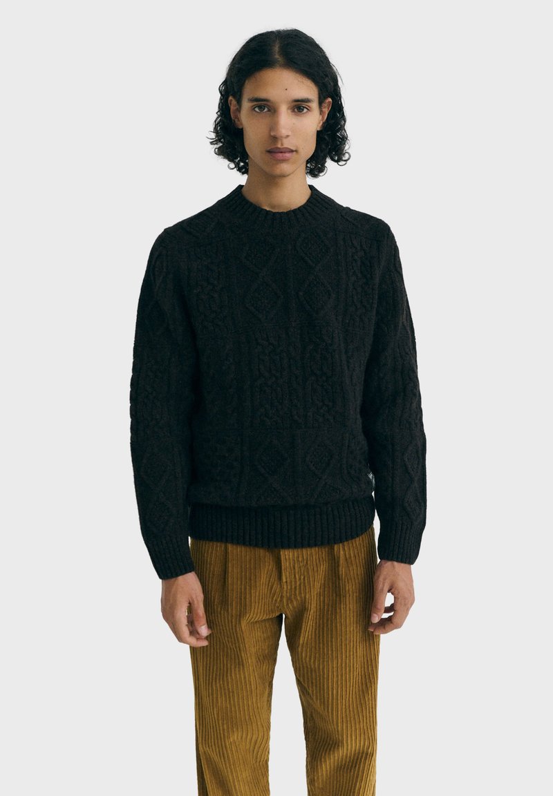 Jersey de punto negro con patrones texturizados, cuello redondo y dobladillo acanalado. Combinado con pantalones de pana de color mostaza.