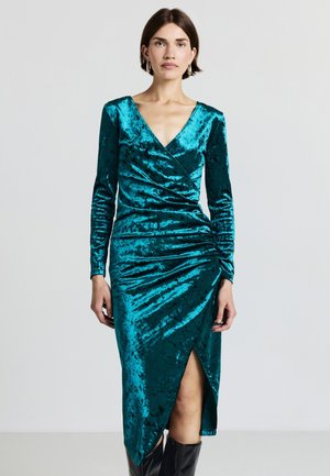 Abito in velluto teal con maniche lunghe, scollo a V e arricciature sul lato. Presenta uno spacco alto fino alla coscia e una silouette aderente.