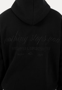 Zwarte hoodie met geborduurde tekst op de achterkant: "niets houdt je tegen", "ESSENTIAL SPORTSWEAR" en een kleine veerafbeelding met cijfers en een copyrightsymbool.