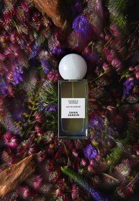 Szklana butelka perfum z białą, kulistą zakrętką, oznaczona etykietami "Vanilla Nomad" i "Sana Jardin", otoczona kolorowymi kwiatami i drewnianymi elementami.