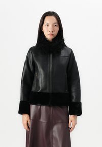 Veste en cuir noir avec un col et des poignets en fourrure, dotée d'une fermeture éclair à l'avant et de deux poches latérales, associée à une jupe en cuir bordeaux.