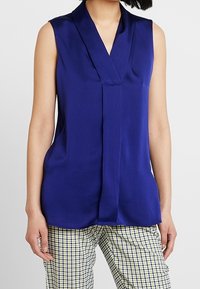 Hedvábná blůza bez rukávů v hluboké modré barvě s výstřihem do V a volným střihem, která má hladký povrch a jemný lesk, spárovaná s kostkovanými kalhotami.