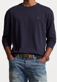 Laivastonsininen pitkähihainen paita pehmeästä kankaasta, pyöreällä kaula-aukolla ja pienellä punaisella logolla rinnassa, yhdistetty haalistuneisiin sinisiin farkkuihin ja ruskeaan vyöhön.