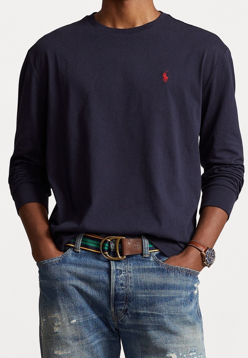 Laivastonsininen pitkähihainen paita pehmeästä kankaasta, pyöreällä kaula-aukolla ja pienellä punaisella logolla rinnassa, yhdistetty haalistuneisiin sinisiin farkkuihin ja ruskeaan vyöhön.