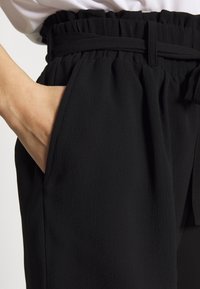 Pantalon noir en tissu texturé, taille élastique et ceinture nouée. Dispose de poches latérales et d'une coupe ample et décontractée.