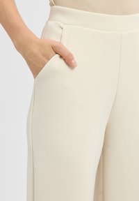 Beige wijde broek met een gladde textuur, uitgerust met zijzakken en een aansluitende tailleband. Focus op strakke lijnen en minimale details.